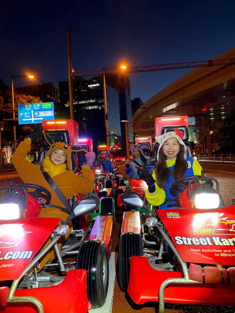 IMG_0022 - StreetKart Group of people in bright costumes posing in red street-kart vehicles on a city street at night, smiling at the camera.