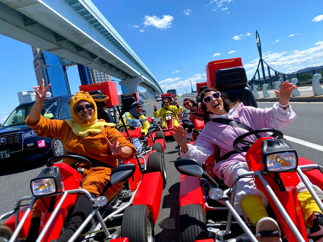 Group of people in bright costumes riding red go-karts on a city road, smiling and waving at the camera under a large bridge.