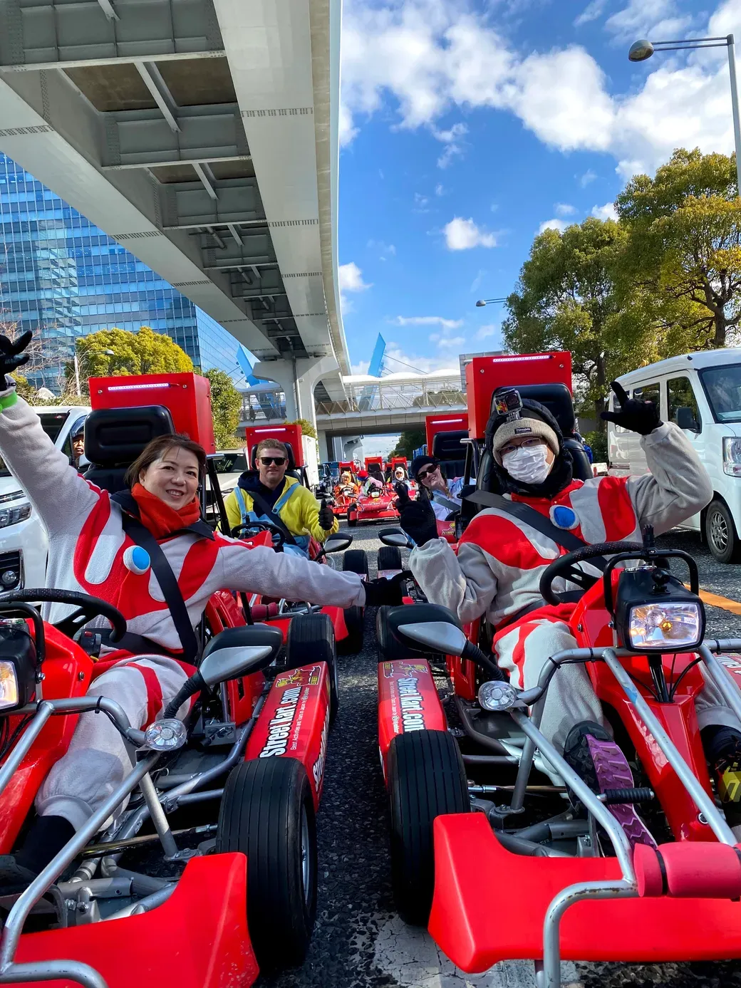 Group of people in red go-karts on a city street under an elevated highway, waving at the camera.