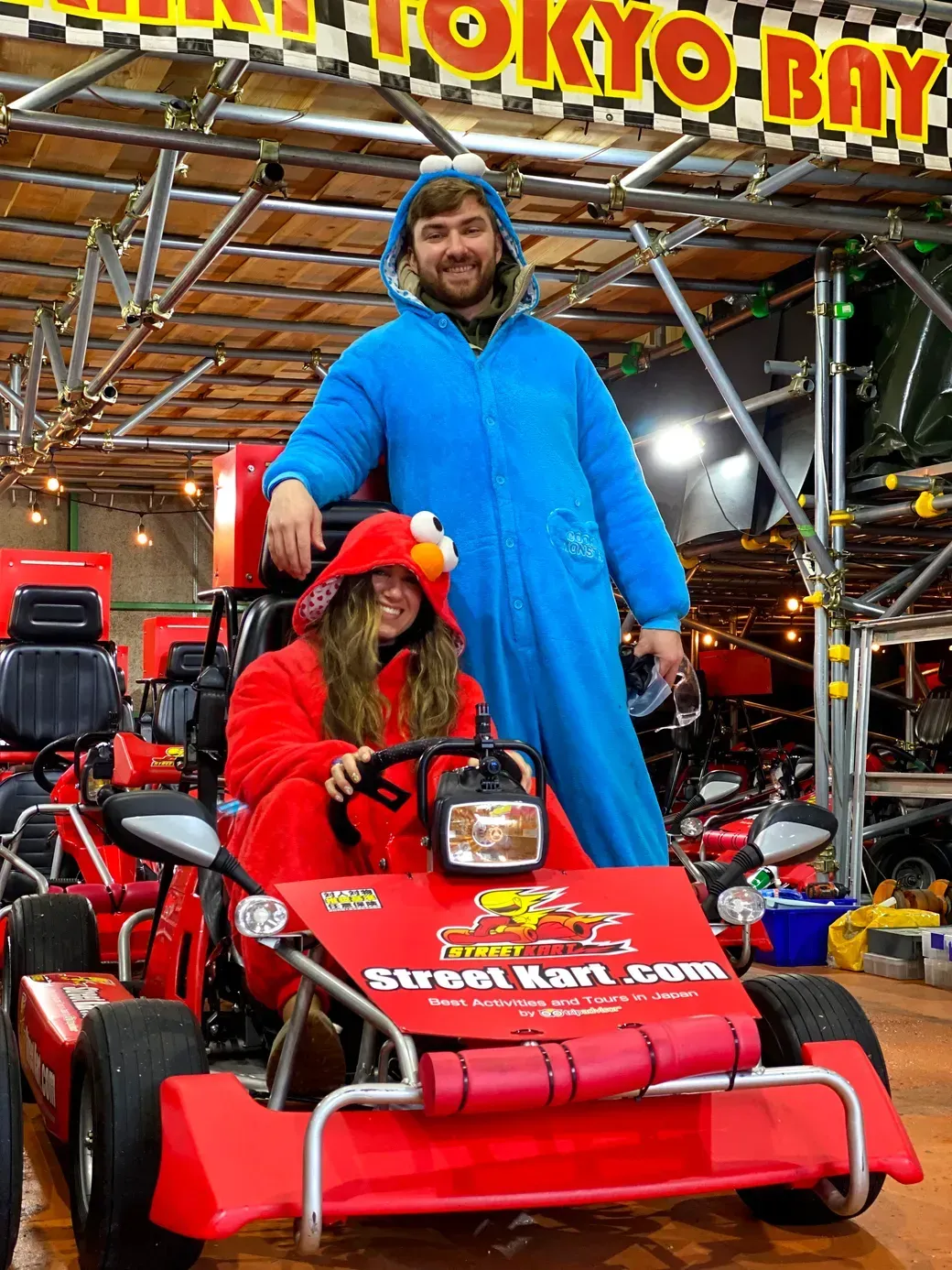 Smiling man in a blue onesie stands behind a red Go-Kart with a woman in a red Elmo costume seated and driving indoors. Signs read Street Kart and Tokyo Bay in the background.